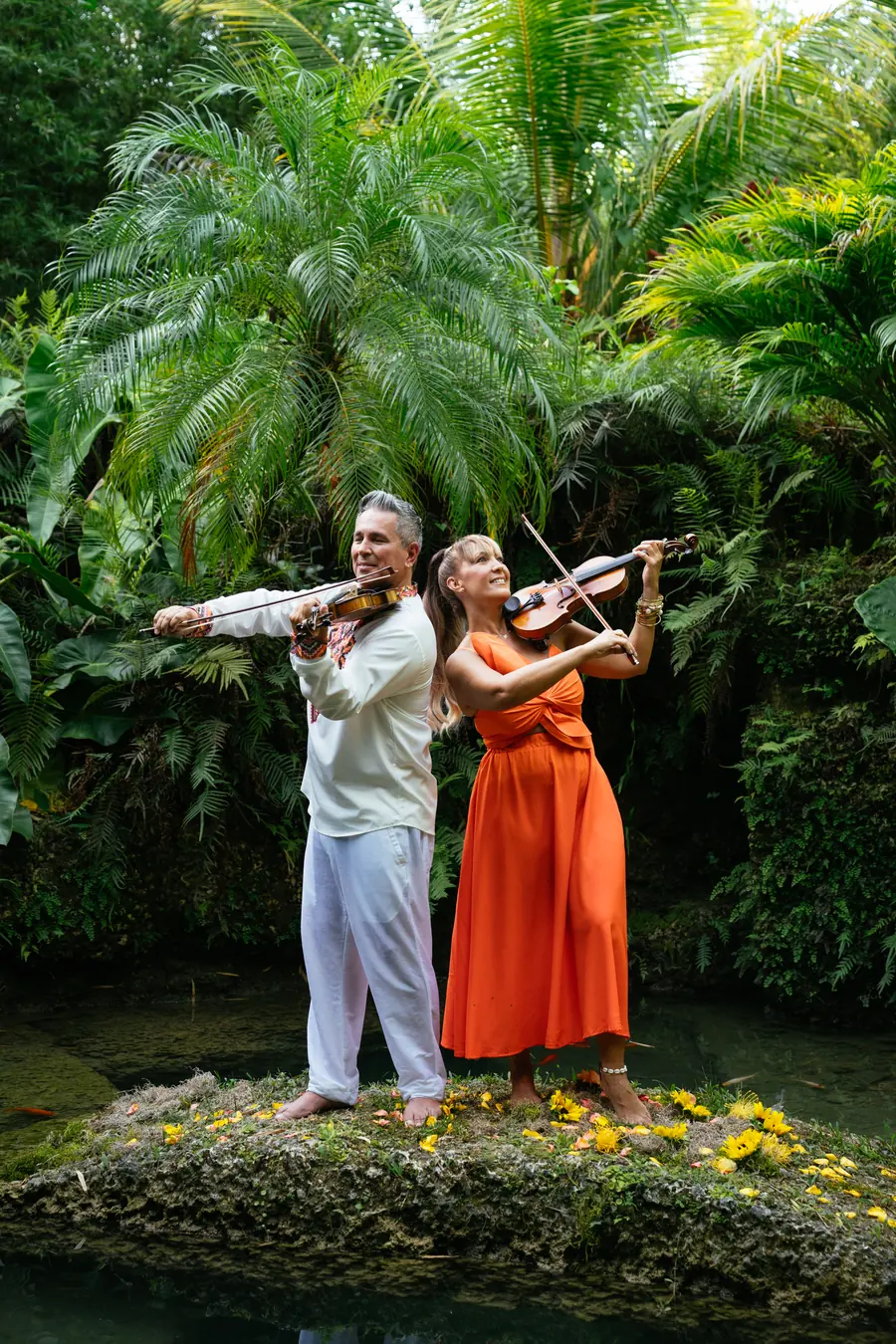 Tania and Abraham raising their violins on a rock in a pond with yellow flowers